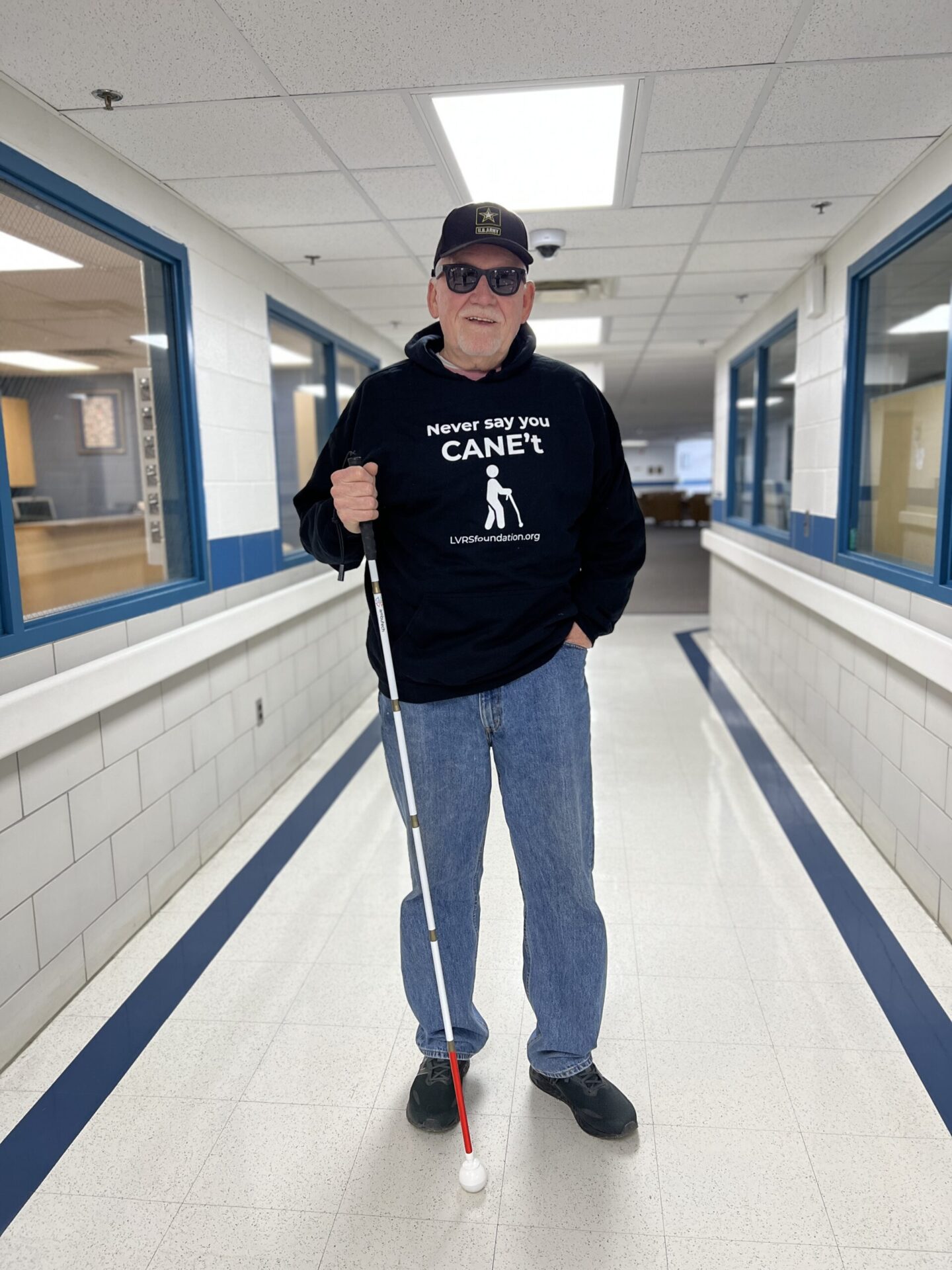 A person wearing a black sweatshirt with the phrase "Never say you CAN'T" stands confidently in a well-lit hallway, holding a white cane.