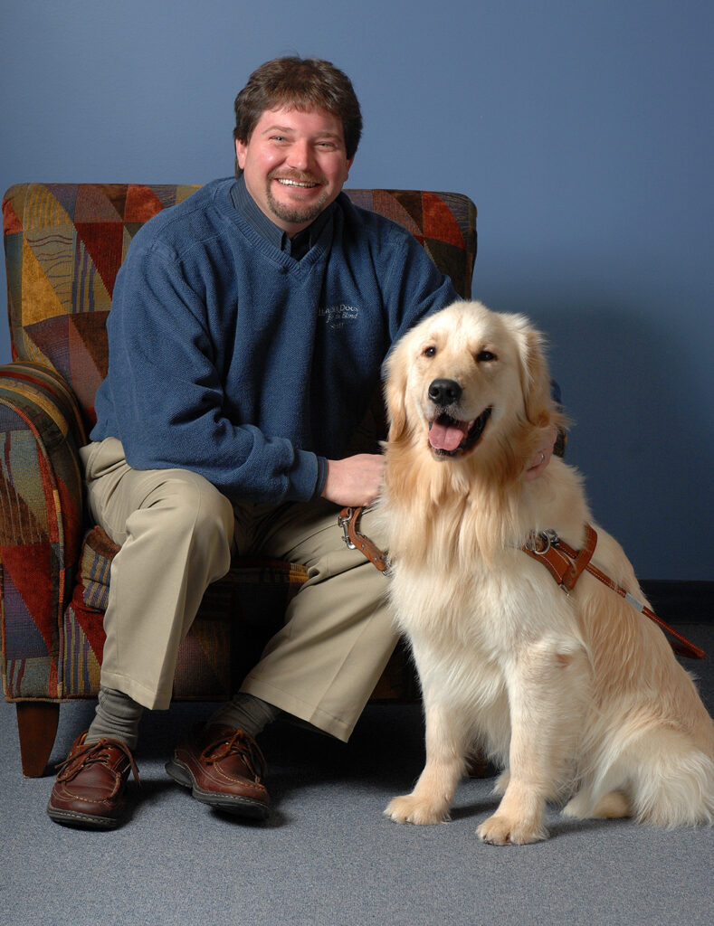 A man sits comfortably in a patterned chair, smiling beside a friendly golden retriever that is wearing a harness.