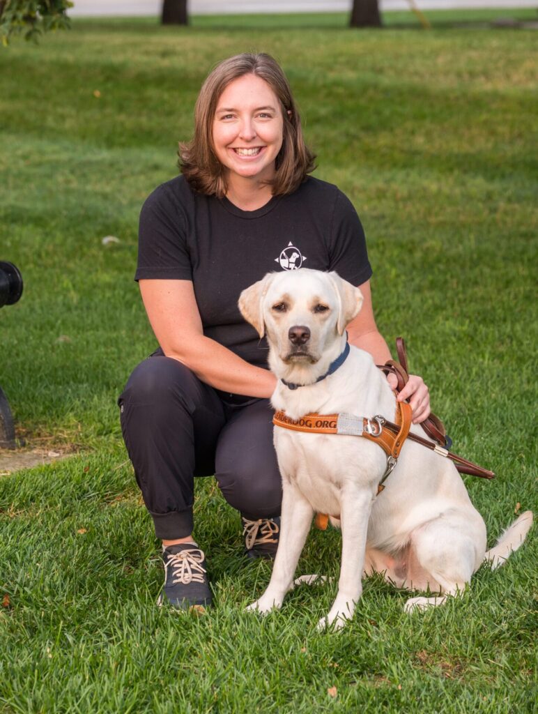 A person with a friendly smile kneels beside a yellow Labrador wearing a harness, set against a grassy backdrop.