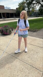 A woman with blond hair uses her long white cane as she walks across the quad. She’s wearing a t-shirt and shorts, and the Fine Arts building can be seen in the background.