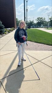 A woman with light blond hair walks down a ramp on campus while using her cane. She is holding her long white cane in one hand and a drink in the other. She’s wearing a hoodie and sweatpants.