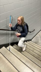 A woman with light blond hair climbs the stairs while holding her drink in one hand and her long white cane in the other. She’s wearing a blue hoodie and gray sweat pants.
