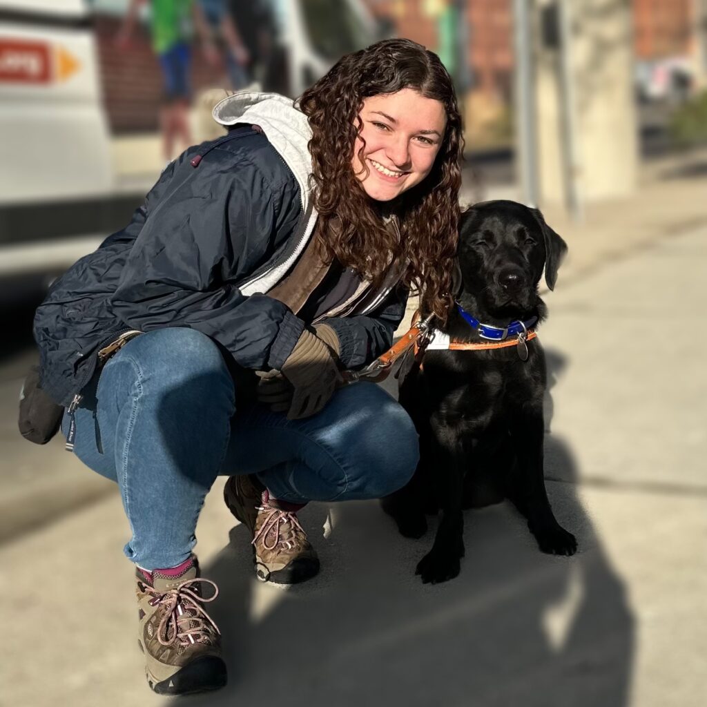 A woman with long brown curly hair smiles toward the camera. She is kneeling next to a black lab Leader Dog in harness.