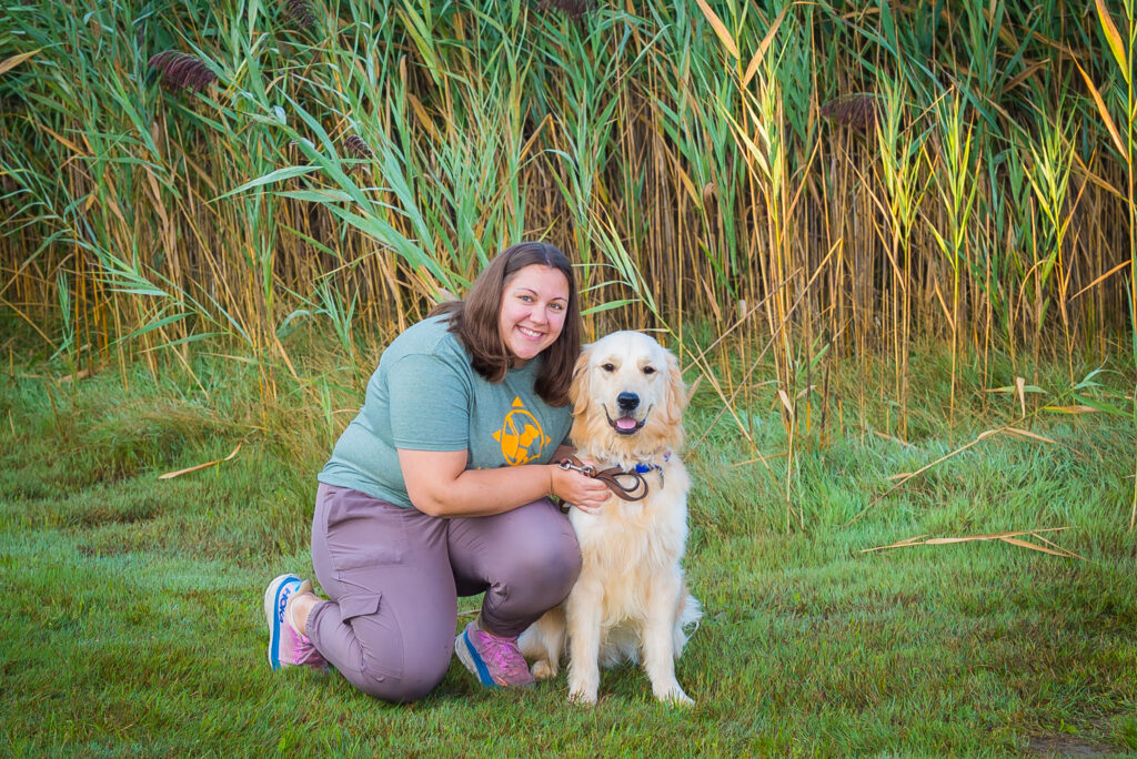 A person kneels beside a golden retriever in a lush, grassy area with tall reeds in the background.