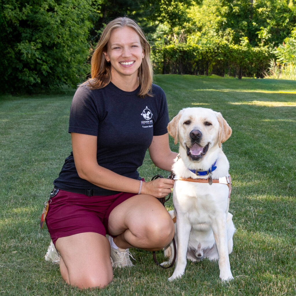 A woman with short blond hair kneels next to a yellow Labrador Leader Dog in a grassy outdoor area.
