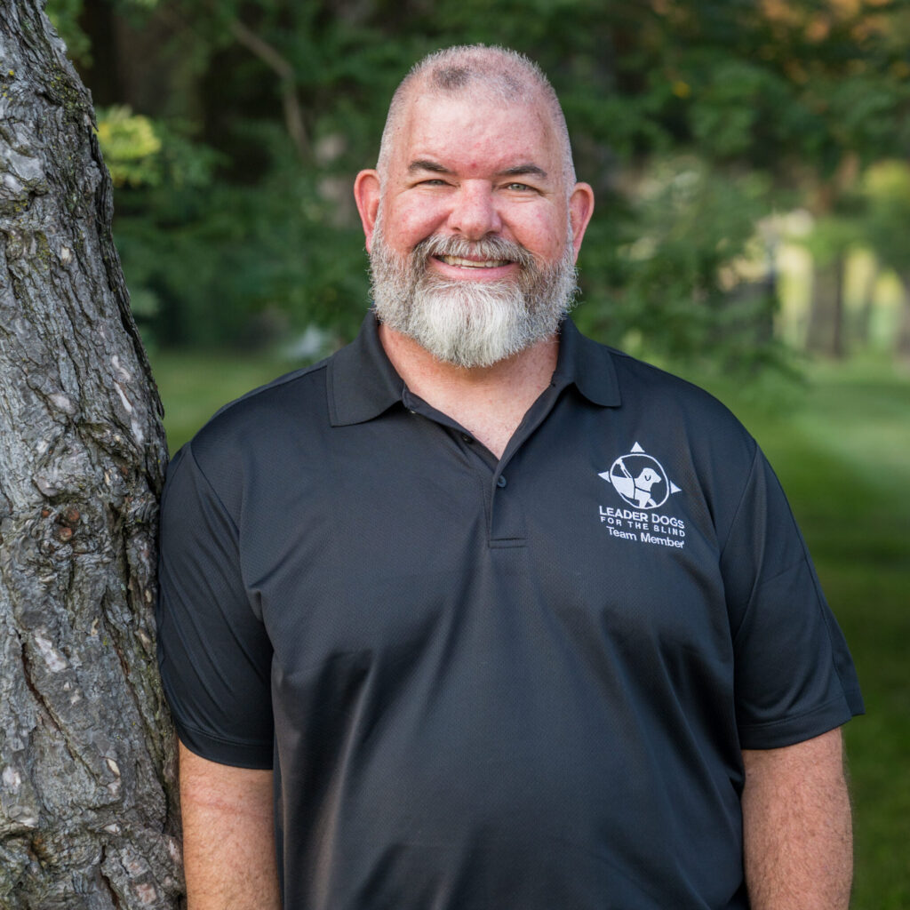 A smiling man with a white beard stands beside a tree, wearing a black polo shirt with a logo.