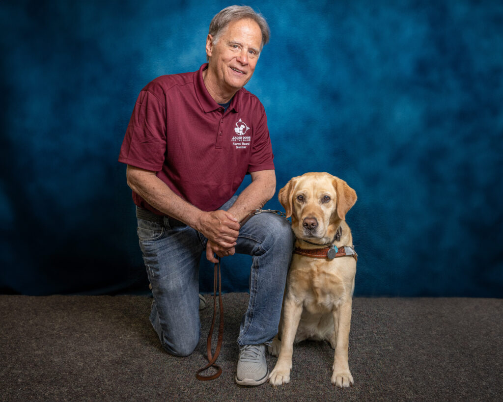 A man smiles toward the camera as he kneels next to his yellow Lab Leader Dog. The dog is in harness and is looking toward the camera.