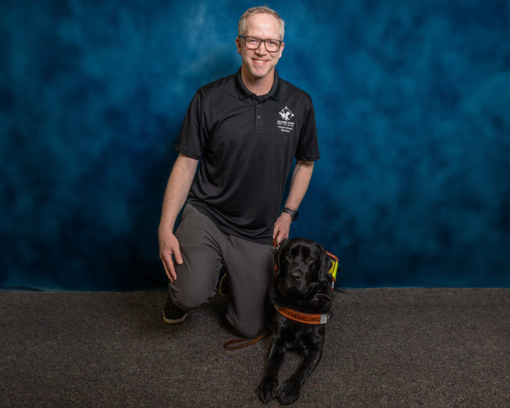 A man with short blond hair smiles toward the camera as he kneels next to his black Lab Leader Dog. The dog is in harness and is also looking toward the camera.