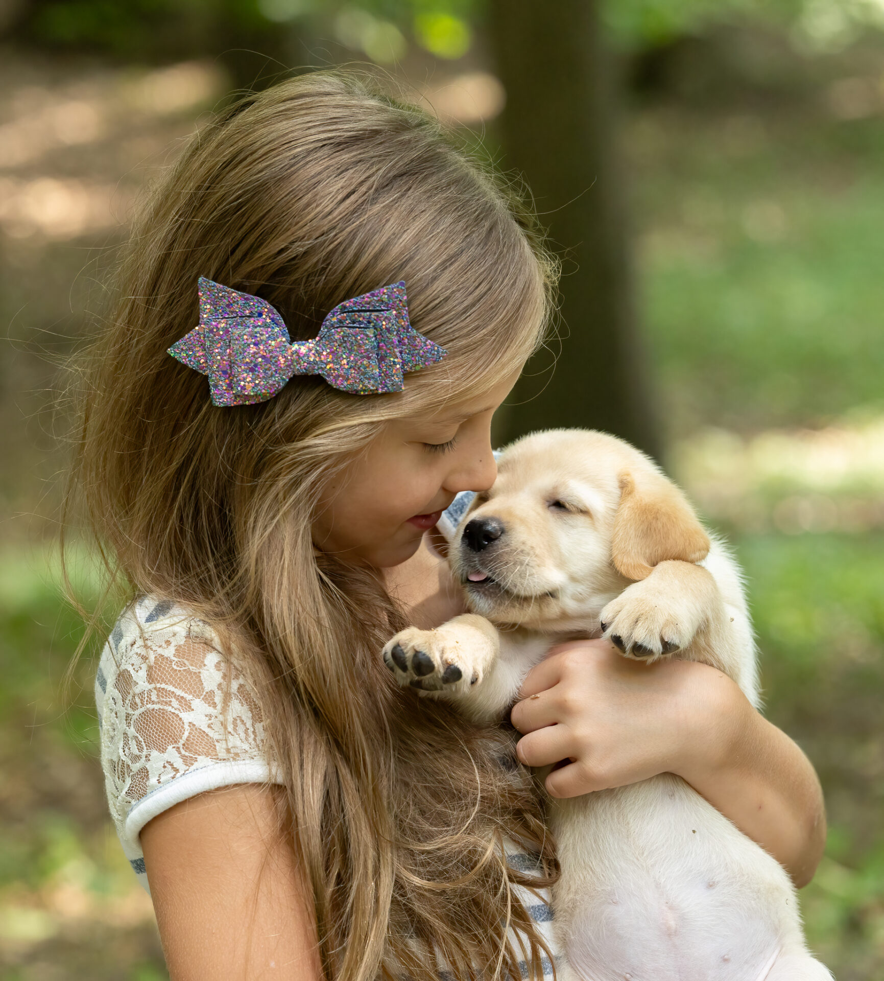 A girl with a sparkly bow in her hair lovingly holds a playful puppy close to her face in a natural, outdoor setting.