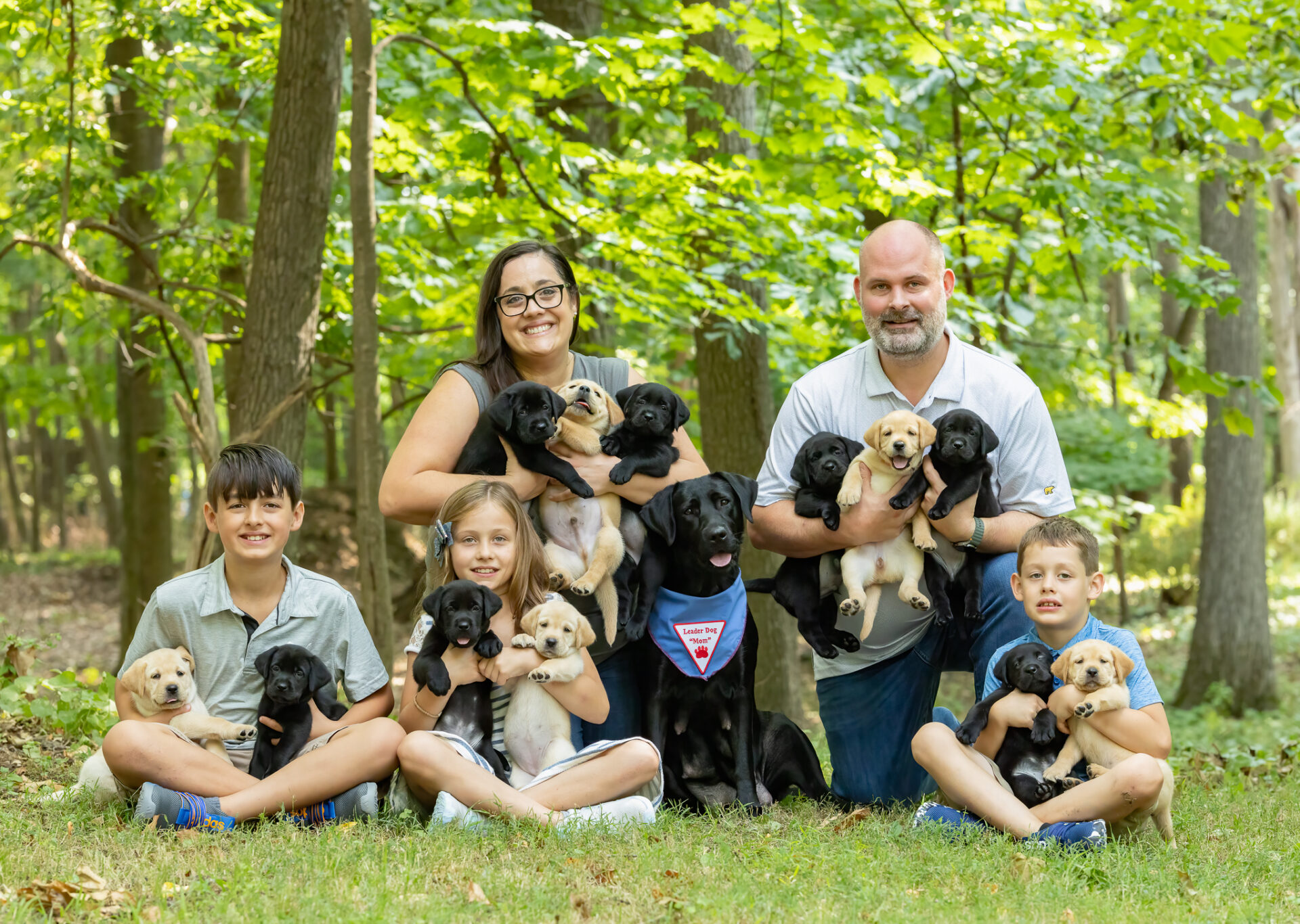 A family poses together in a wooded setting, holding several adorable puppies while smiling for the camera.