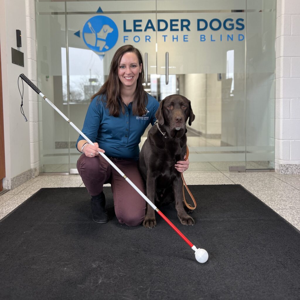 A woman in a blue jacket kneels beside a brown guide dog while holding a white cane, with a logo for Leader Dogs for the Blind in the background.