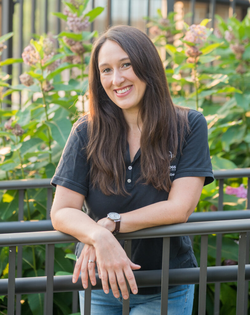 A person with long hair smiles while leaning casually on a railing surrounded by greenery and flowers.