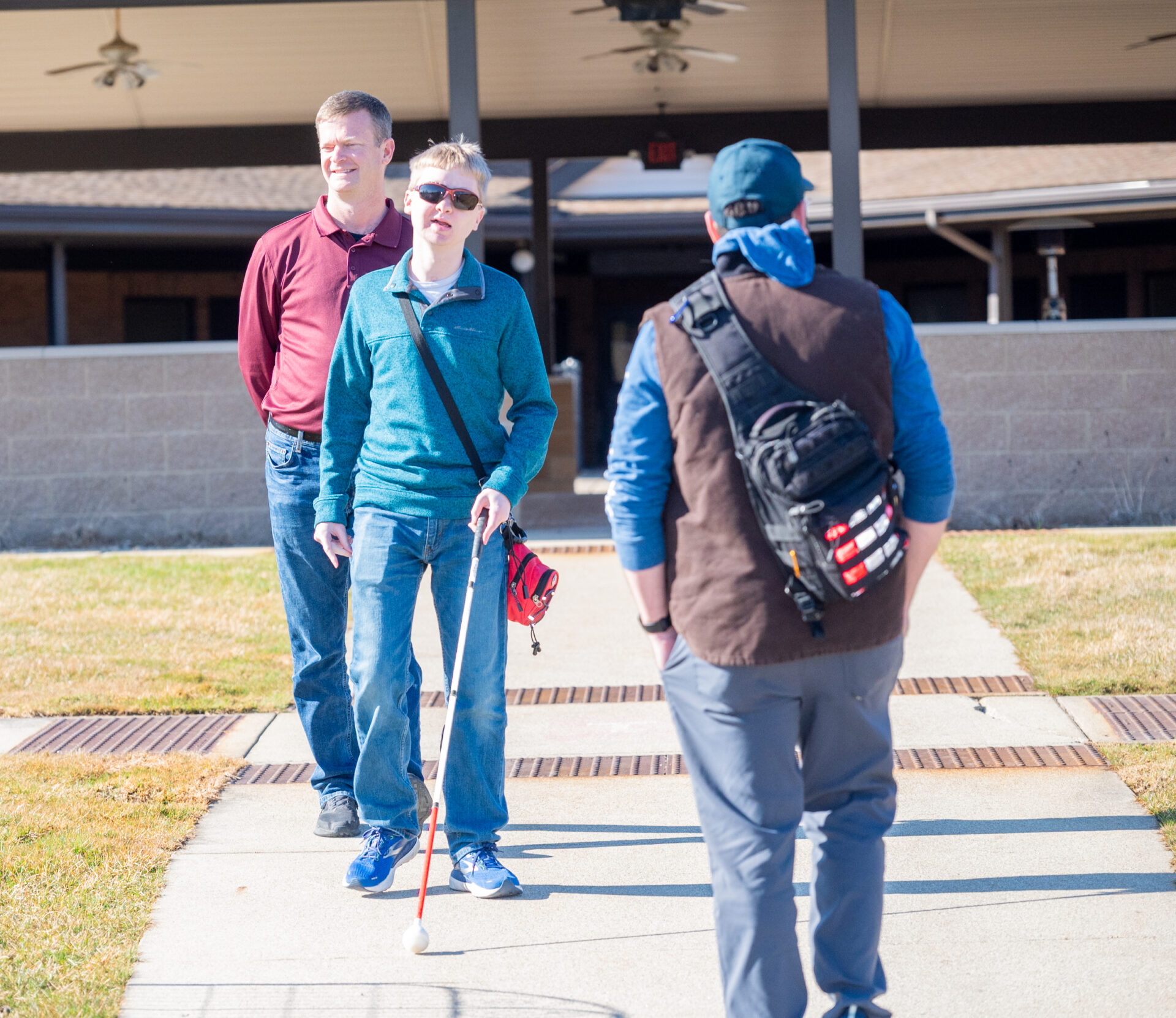 A person with a white cane stands on a sidewalk while two others walk nearby in a sunny outdoor setting.