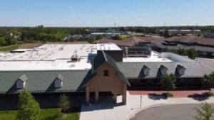 A large building featuring a sloped green roof and a welcoming entrance is surrounded by greenery and other structures in a sunny landscape.