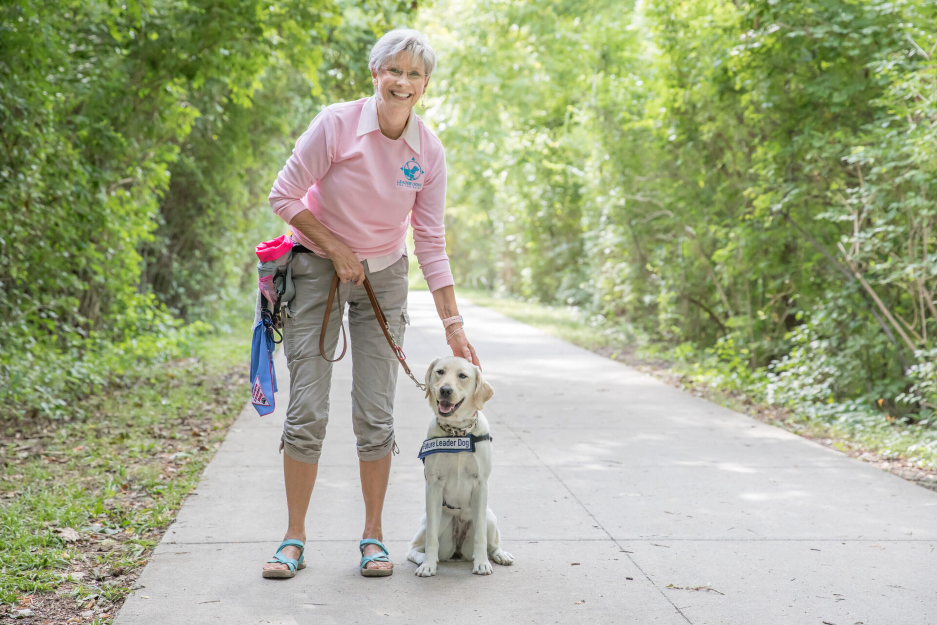 A woman in a pink shirt and shorts stands beside a happy Labrador retriever on a shaded pathway surrounded by greenery.