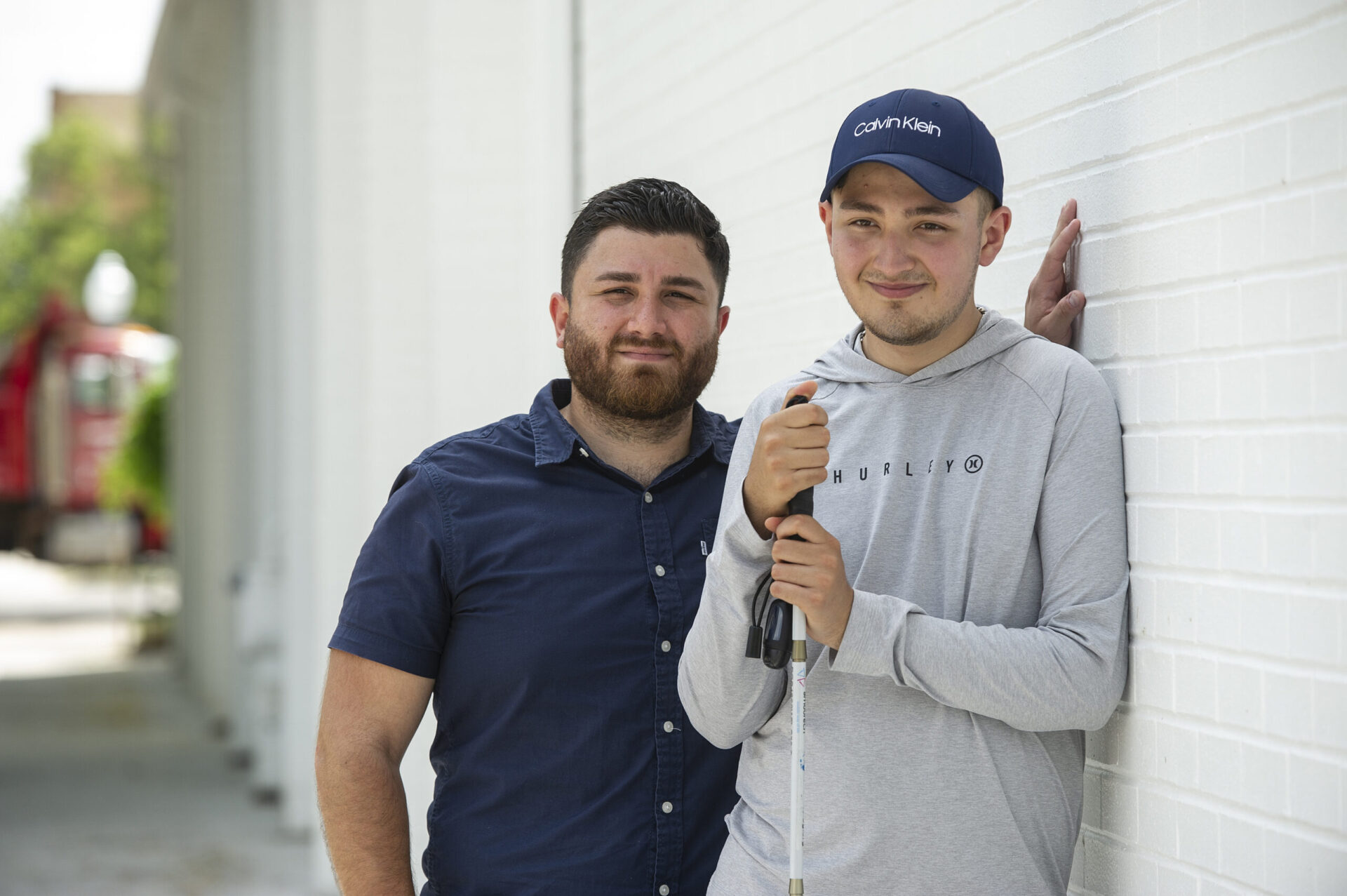 Two smiling men pose together against a white wall; one wears a Hurley hoodie and Calvin Klein cap while holding a white cane.