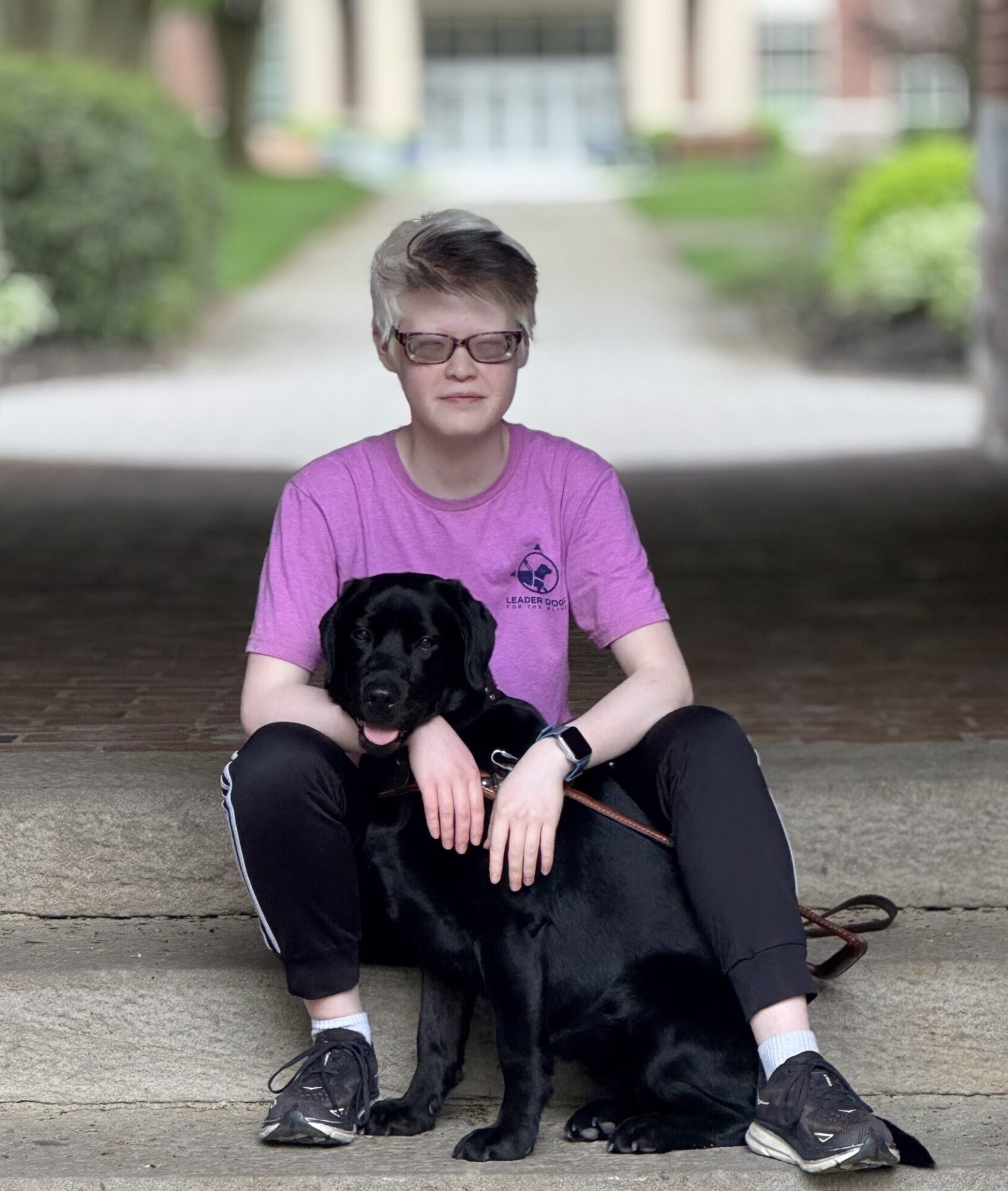 A person in a pink shirt sits on steps with a black dog beside them.