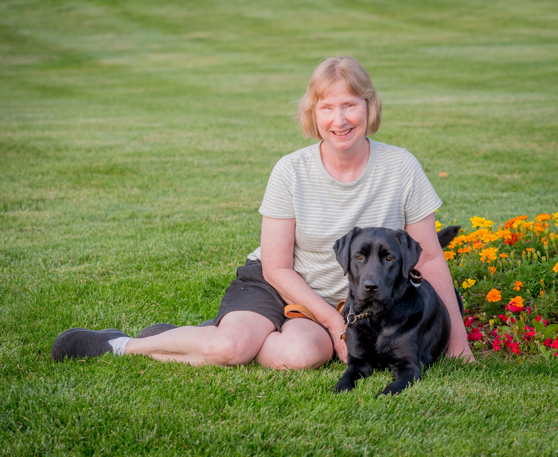 A woman with short blond hair sits on a grassy field. Laying down next to her is a black lab guide dog.