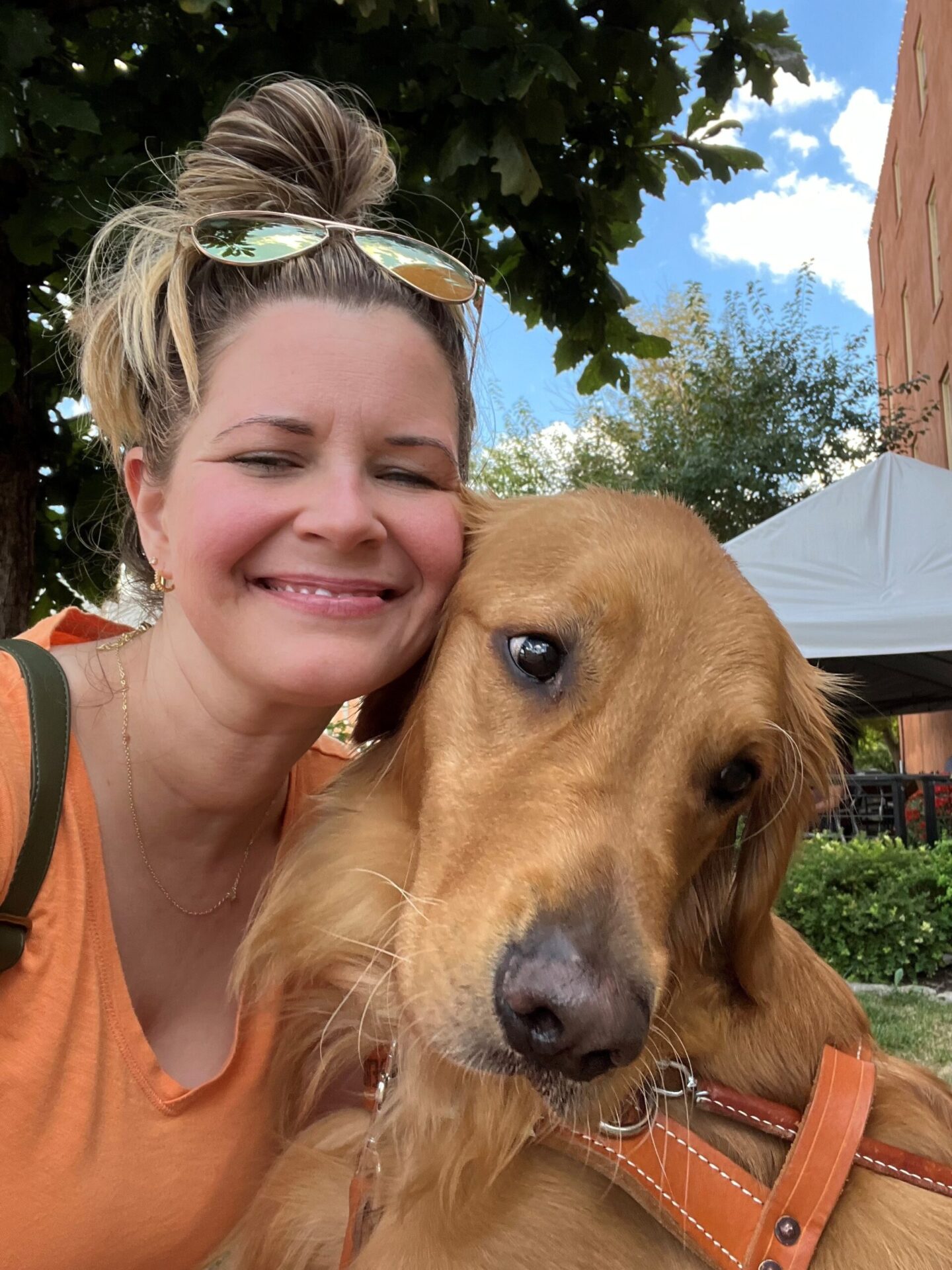 A woman smiles while posing closely with a golden retriever, both enjoying a sunny day outdoors.