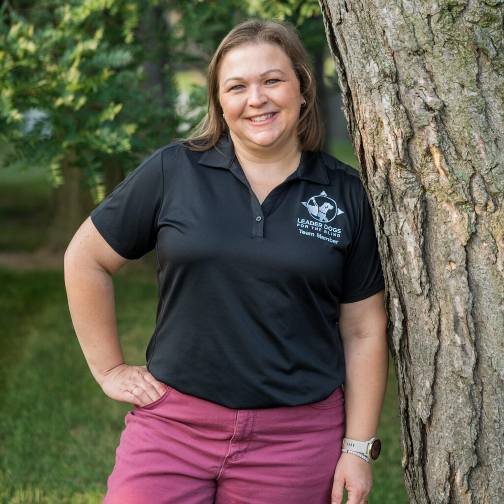 A person stands casually next to a tree, wearing a black polo shirt and pink pants, with a friendly smile.