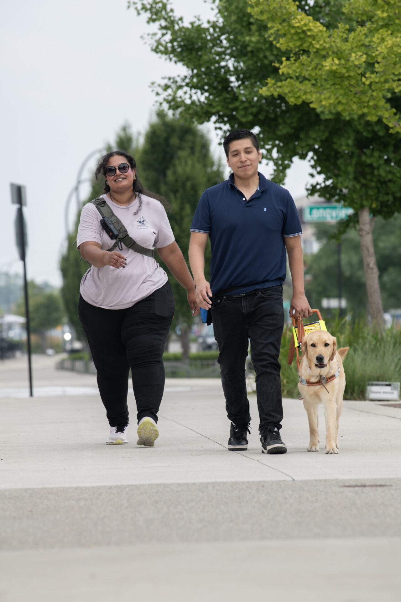 A man walks with a guide dog on a wide sidewalk. A couple of steps behind him is a woman wearing a crossbody pack. They smile as they walk toward the camera.