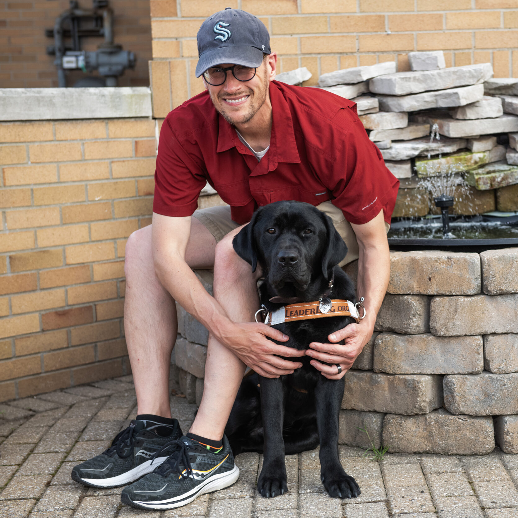 A man sits at the ledge of a small water fountain display. He wears a ball cap and red shirt. He leans forward to hug a black lab guide dog that is sitting at his feet.