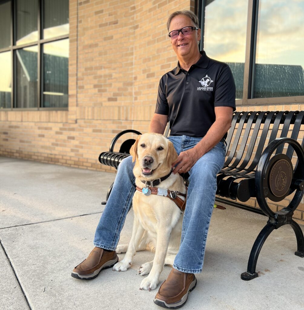 Man in black polo shirt and glasses sitting on decorative black metal bench with yellow Labrador wearing guide dog harness positioned between his legs, in front of brick building with large windows.
