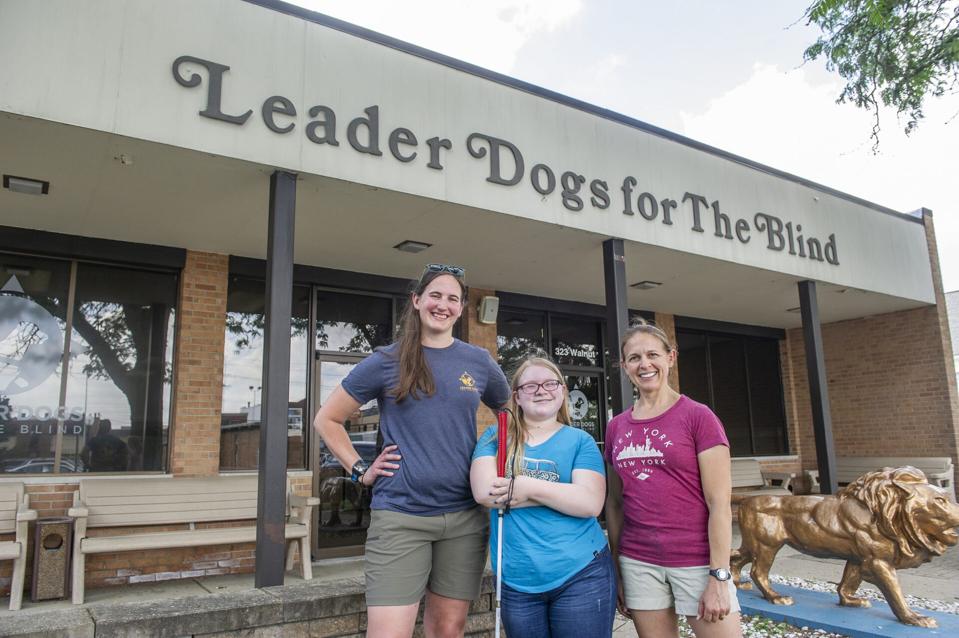 Three smiling women standing outside Leader Dogs for the Blind building, with the center woman holding a white cane and a golden lion statue visible to the right.