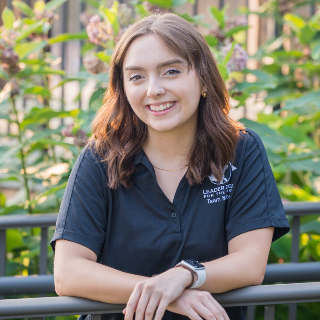 A person with shoulder-length brown hair smiles while leaning against a railing surrounded by lush greenery.