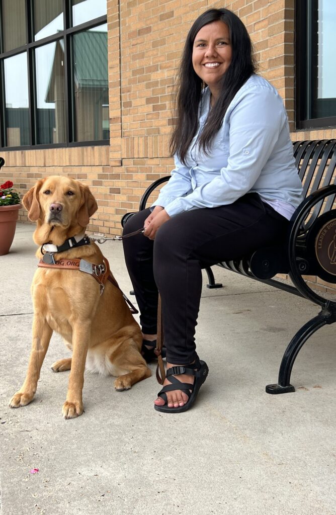 Woman in light blue shirt sitting on decorative black metal bench with golden Labrador wearing guide dog harness sitting attentively beside her, on covered walkway with brick wall, windows, and flower planter in background.