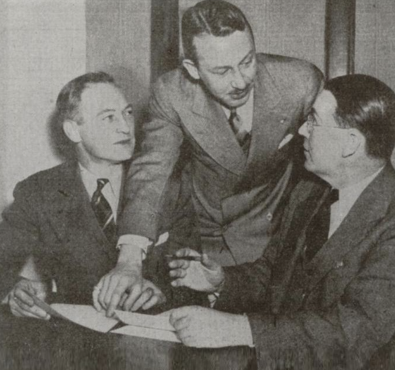 Three men engaged in a discussion, with one leaning in to share a point while the others listen attentively.