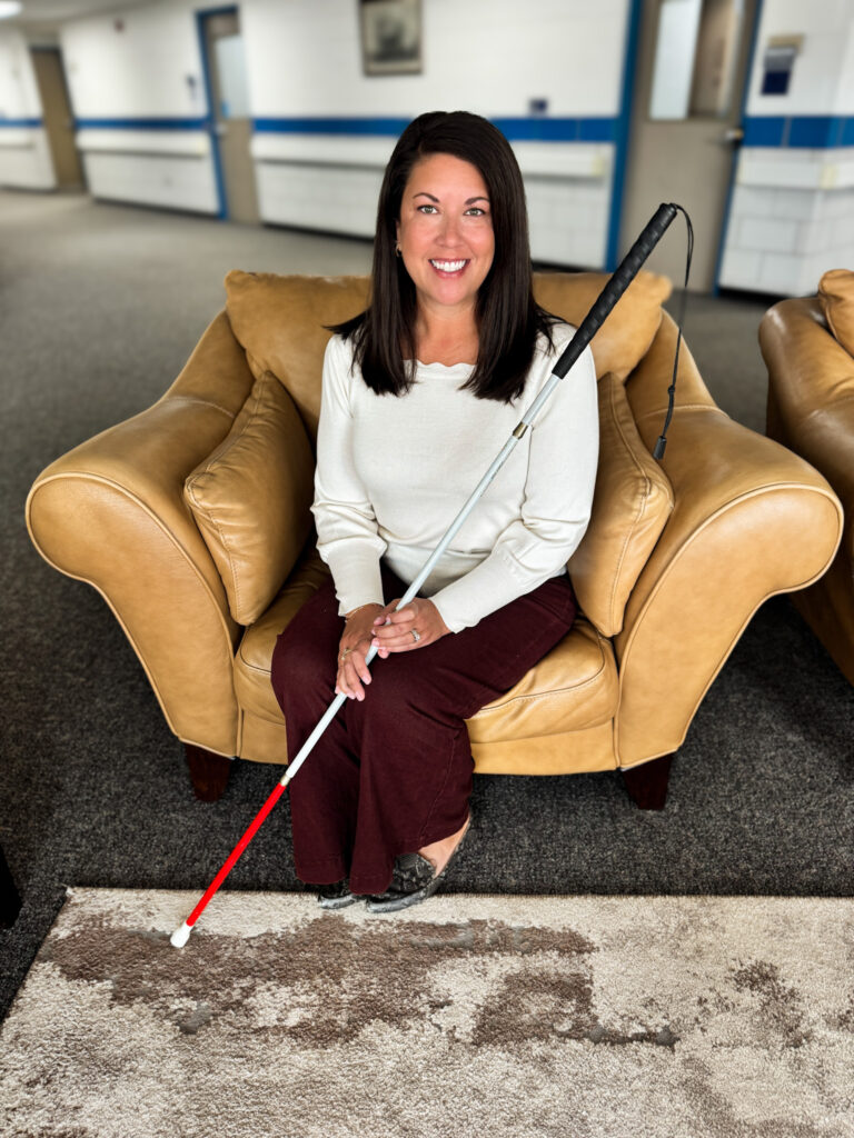 A person sits comfortably on a tan leather couch, holding a white cane and smiling warmly.