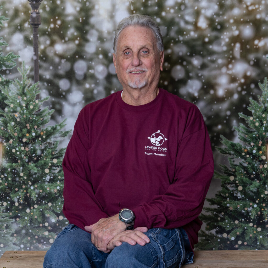 A man in a maroon sweatshirt and blue jeans sits on a wooden bench against a festive backdrop with Christmas trees and glowing lanterns.