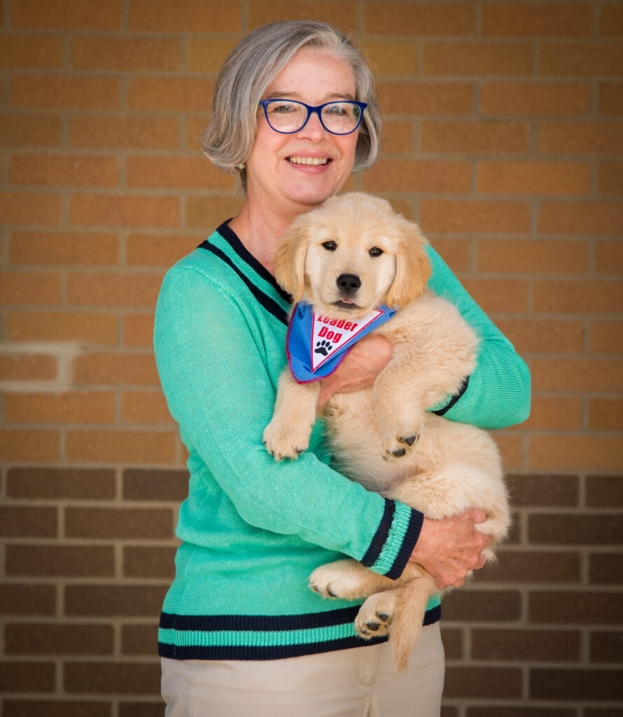 A woman with gray hair wearing glasses smiles while holding a golden retriever puppy in her arms.