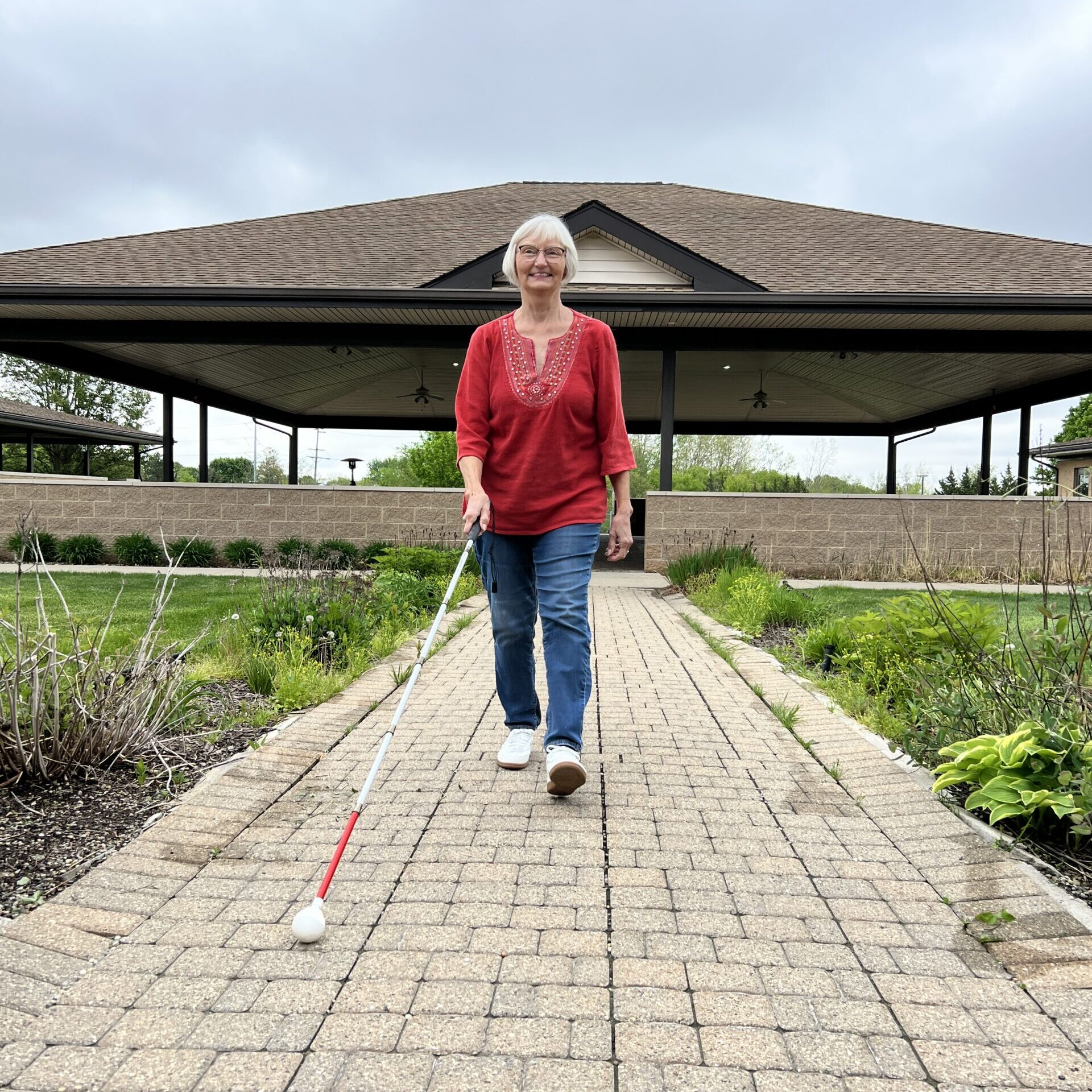A person walks confidently along a paved pathway, using a white cane, with a large pavilion in the background under a cloudy sky.