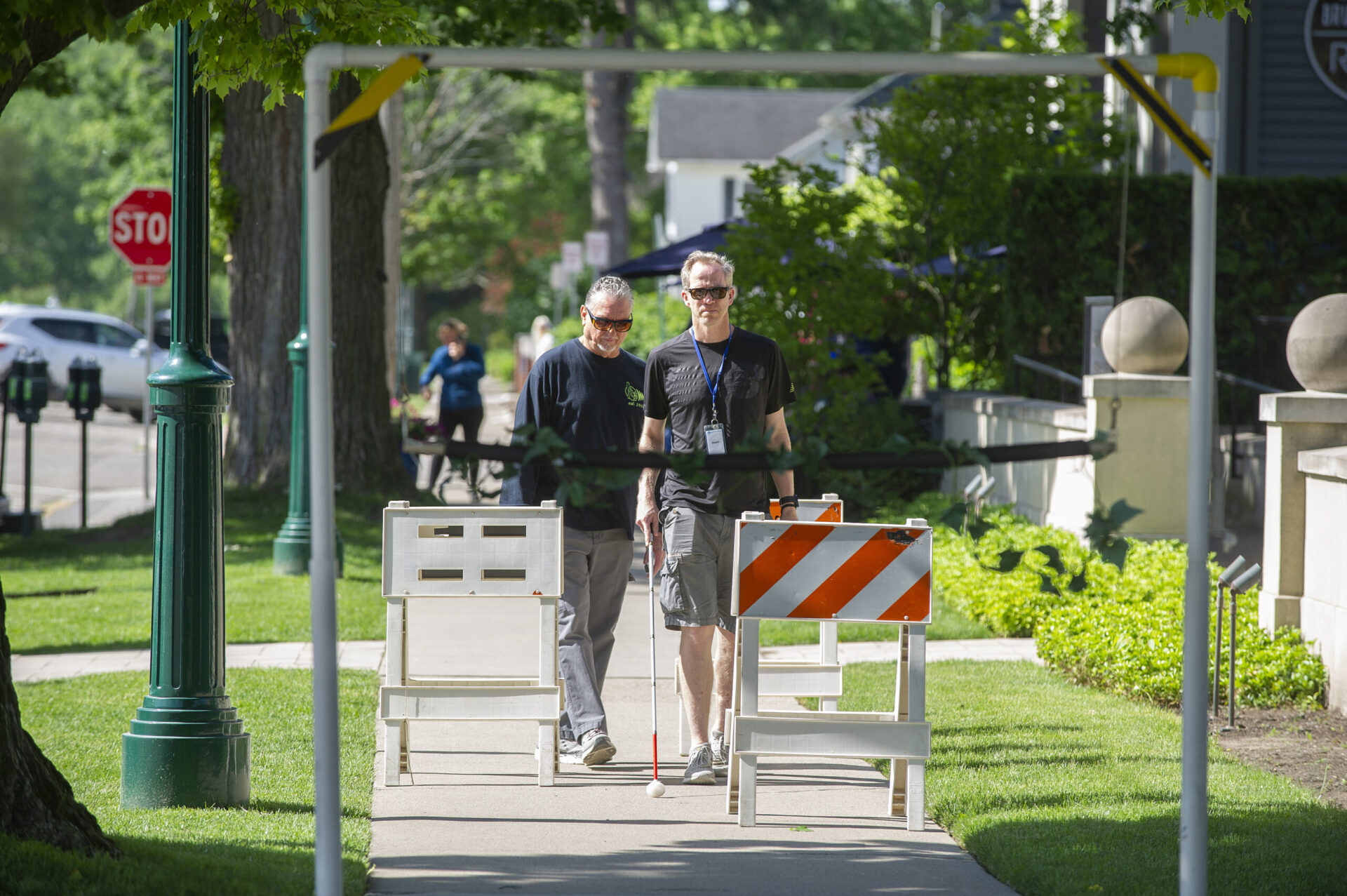 A person is guiding another individual past a construction barrier on a sidewalk lined with trees and greenery.