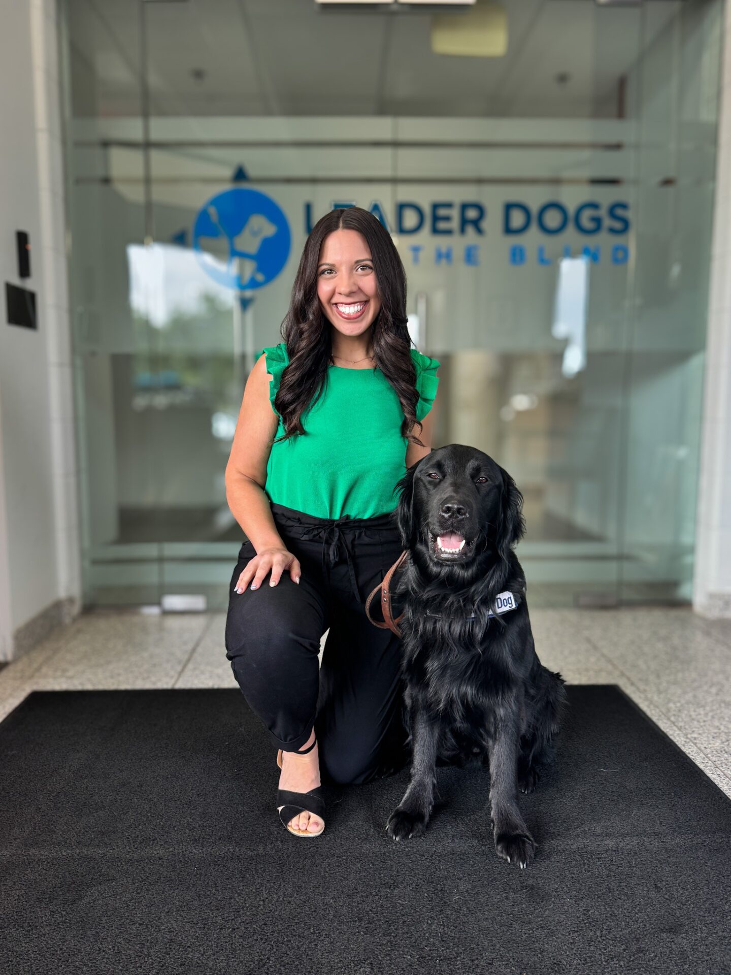 A smiling woman in a green top kneels beside a black dog with a service vest in front of a glass door featuring the "Leader Dogs for the Blind" logo.