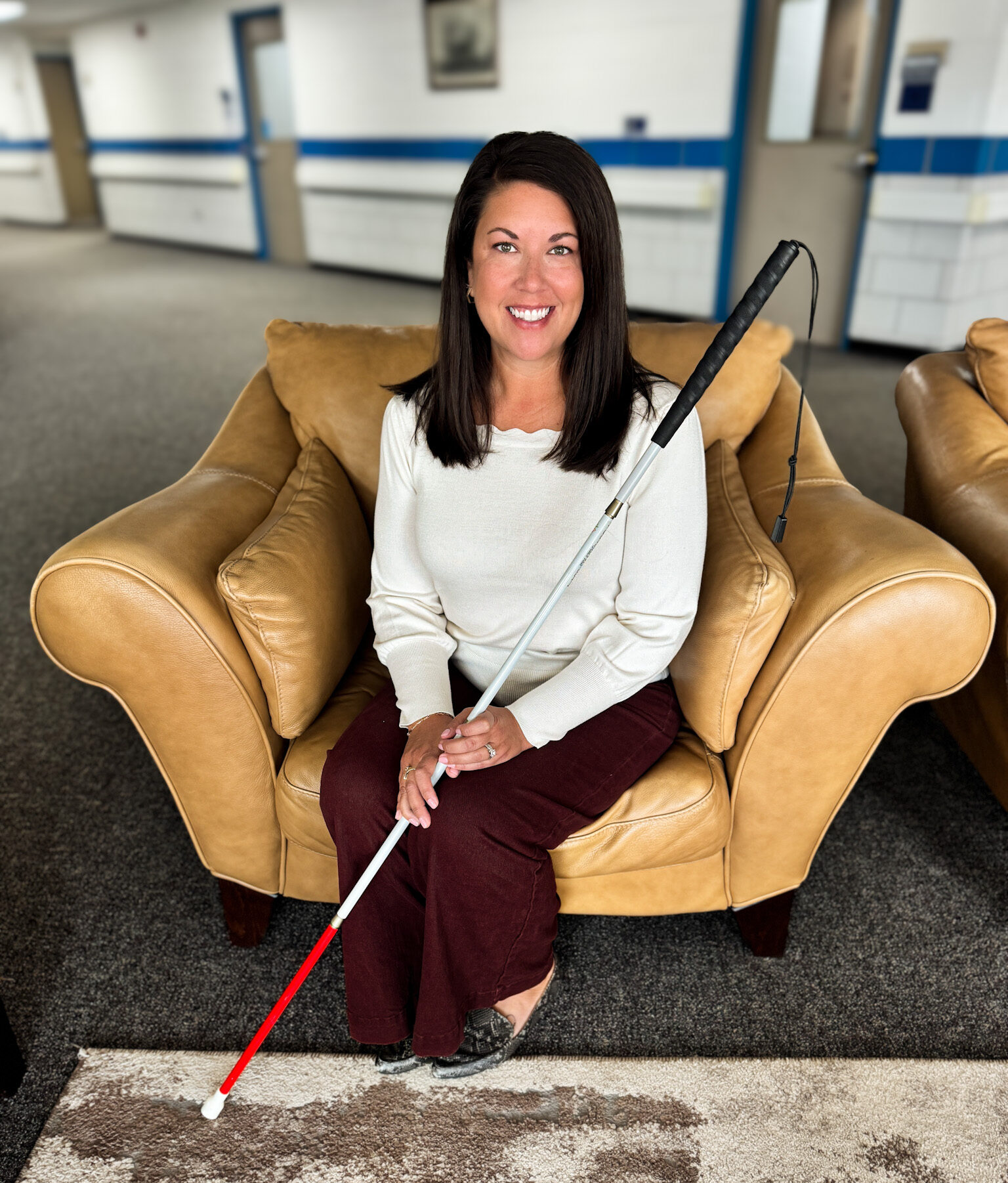 A person sits comfortably on a tan leather couch, holding a white cane while smiling at the camera.