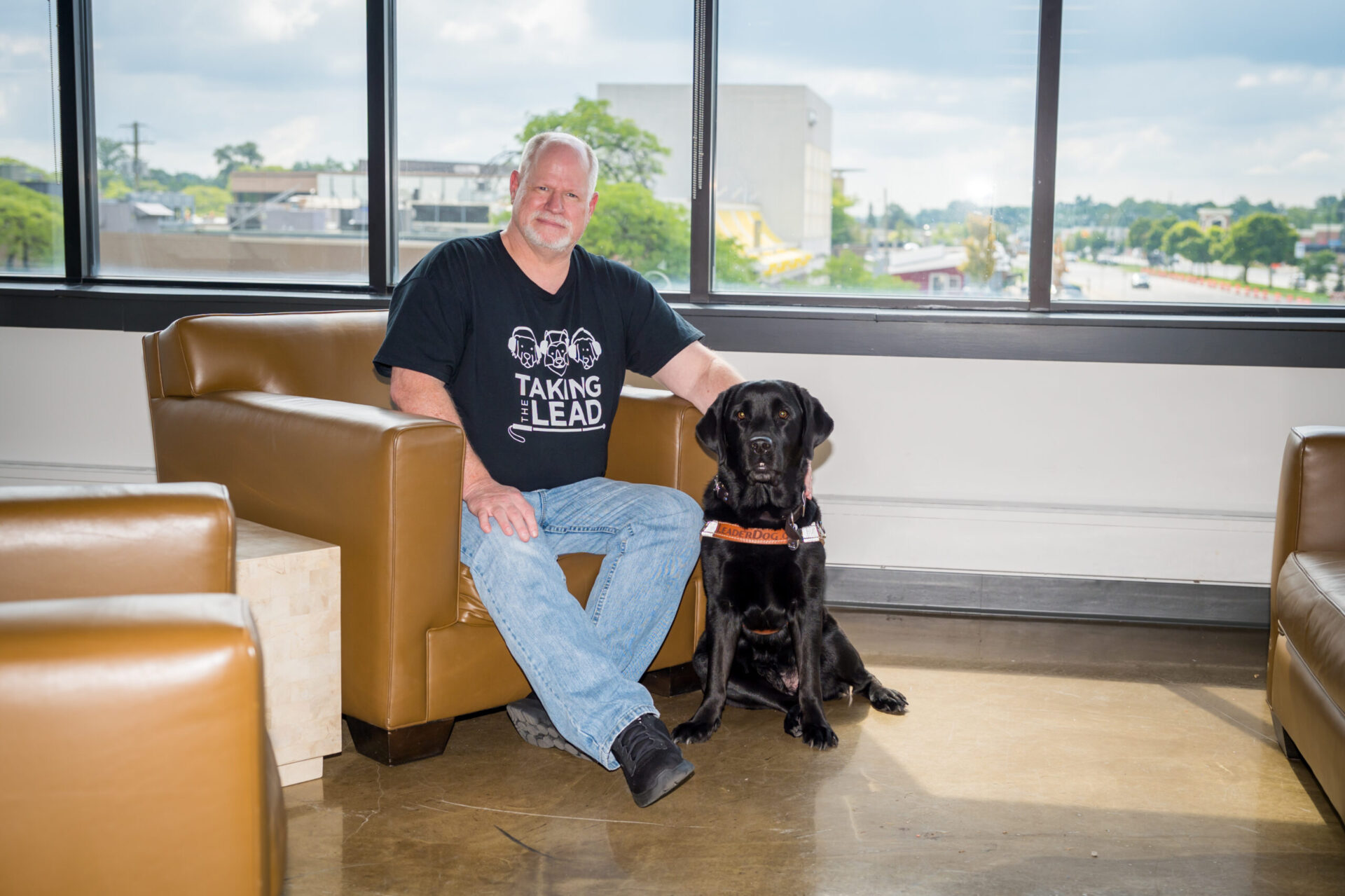 A person wearing a black "Taking the Lead" t-shirt sits comfortably in an armchair beside a black Labrador Retriever in a bright, spacious interior with large windows.