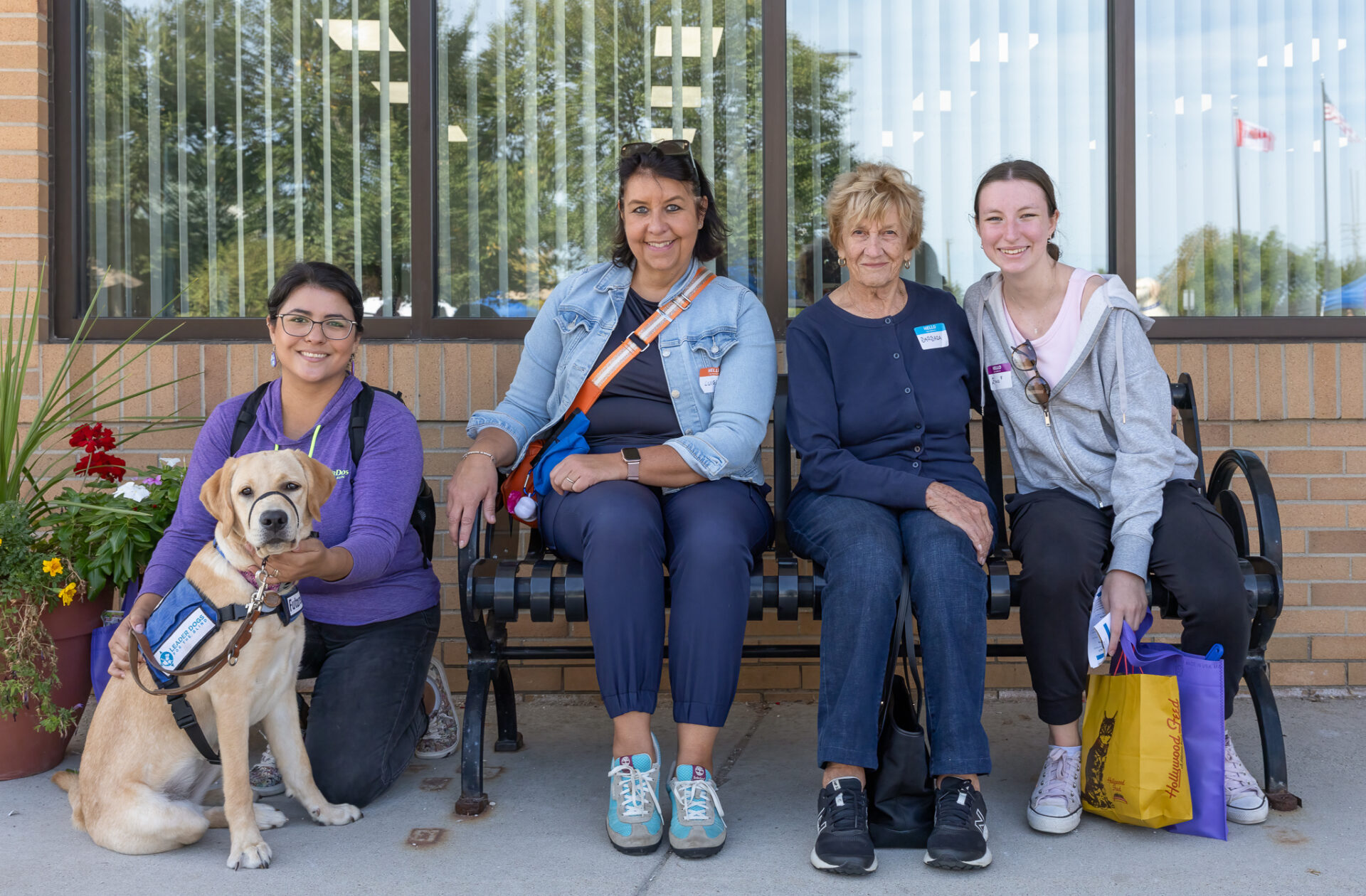 A group of four individuals, including a dog, sits on a bench in front of a building with large windows, smiling and engaging with each other.