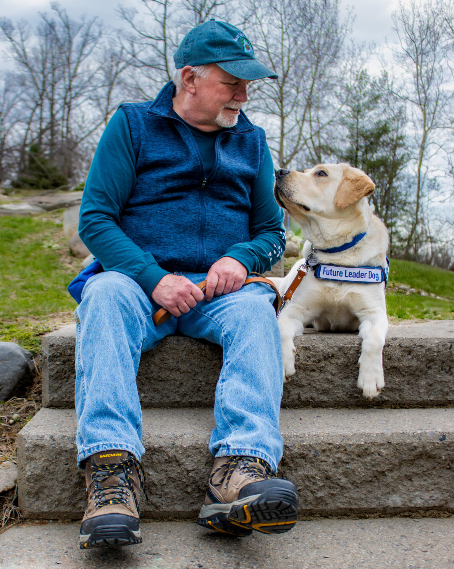 A man sits on a stone step, gazing affectionately at a golden Labrador wearing a harness labeled "Future Leader Dog."