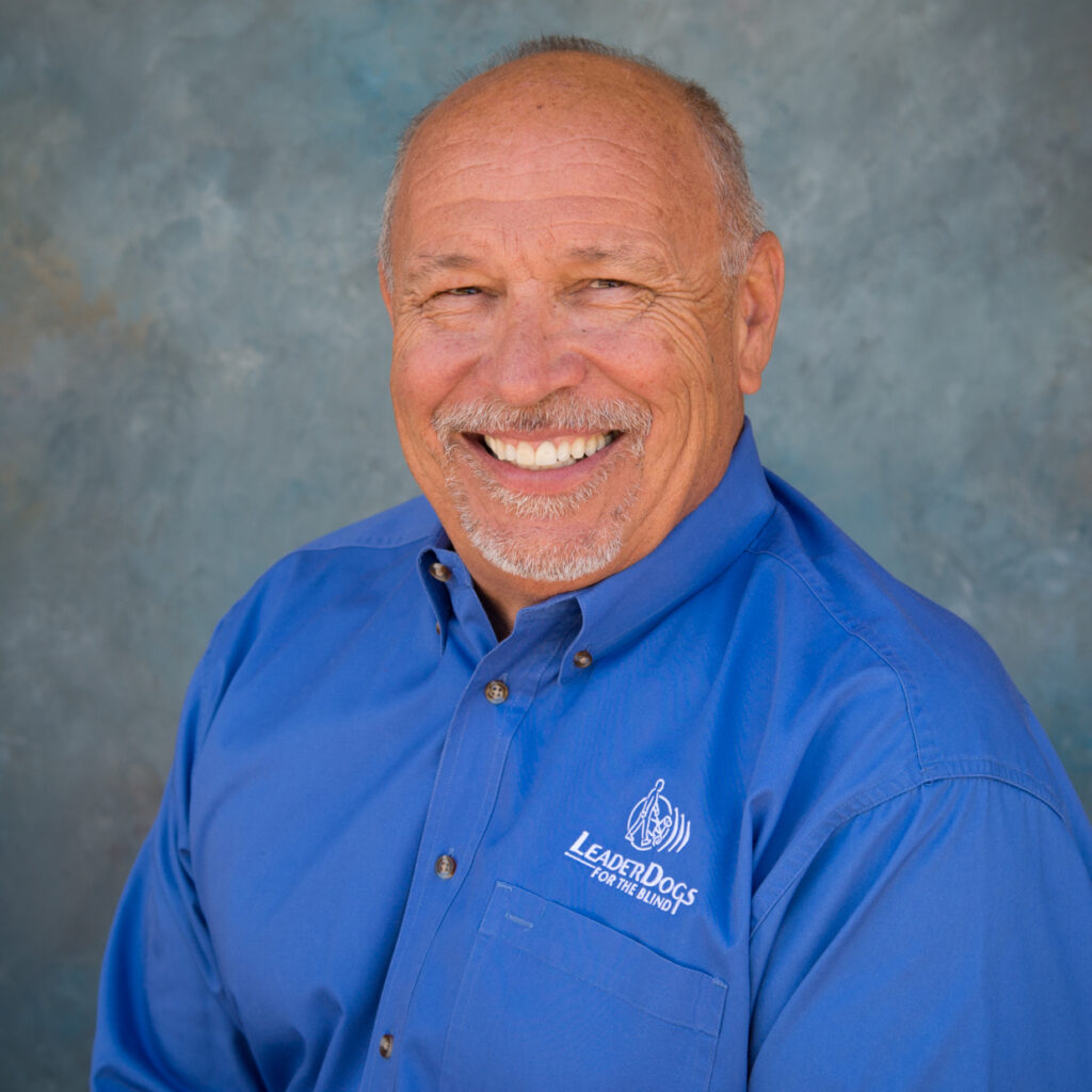 A smiling individual wearing a blue shirt with a logo, set against a textured backdrop.
