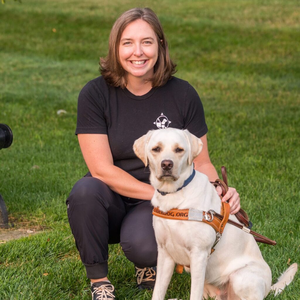 A woman kneels beside a yellow Labrador retriever wearing a harness, both smiling in a grassy outdoor setting.