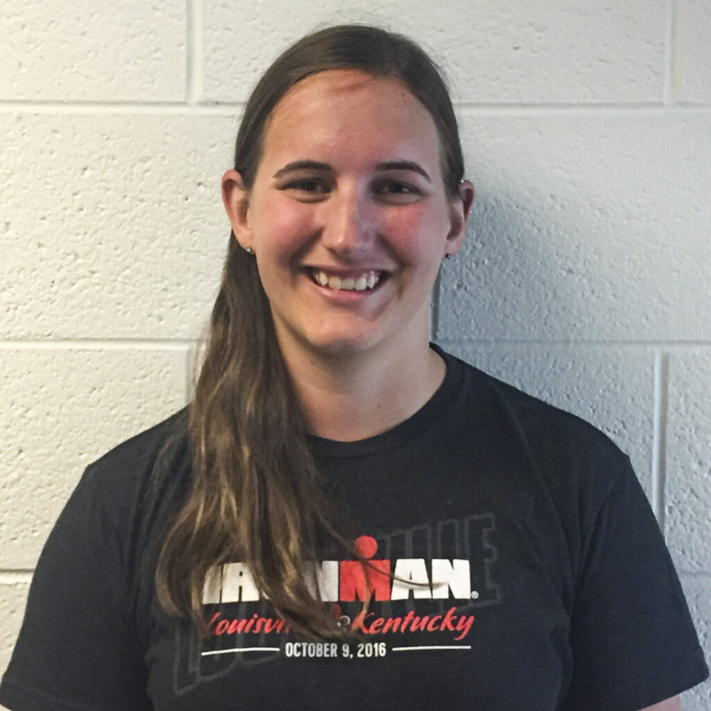 A person with long hair is smiling while wearing a dark t-shirt featuring the words "Ironman" and a date.