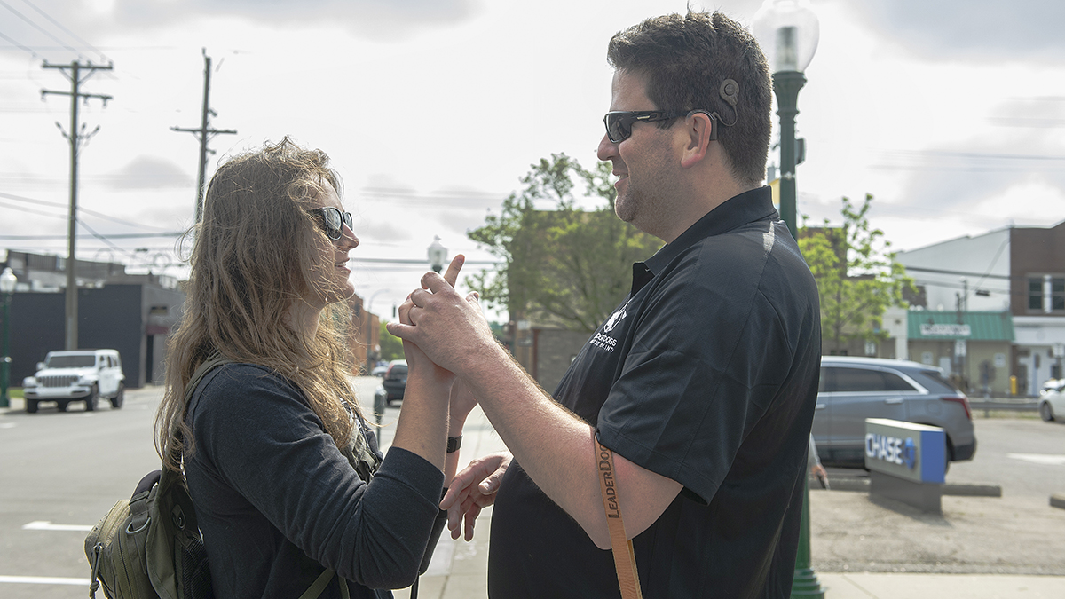 Two people are engaged in a lively conversation on a city street, with one holding a smartphone and both wearing sunglasses.
