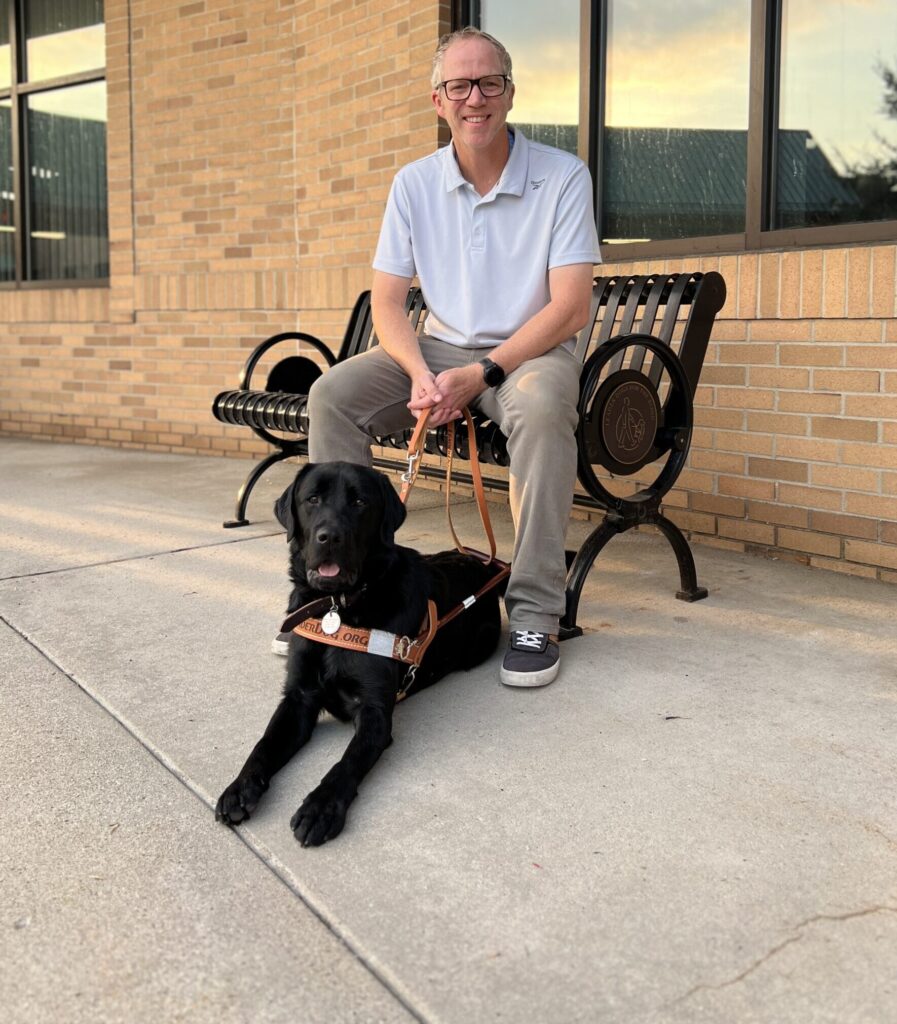 Man sitting on decorative bench with black Labrador guide dog lying at his feet in front of brick building.