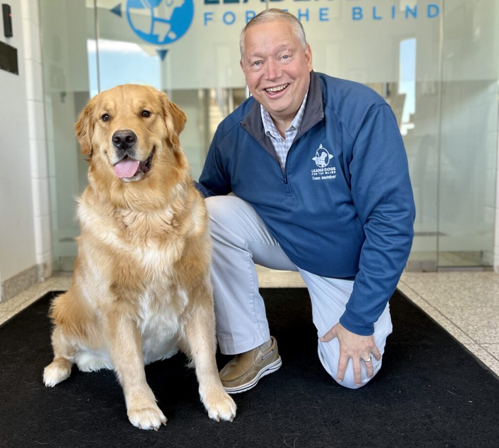 A smiling man in a blue jacket poses alongside a golden retriever on a black mat in a brightly lit office space.