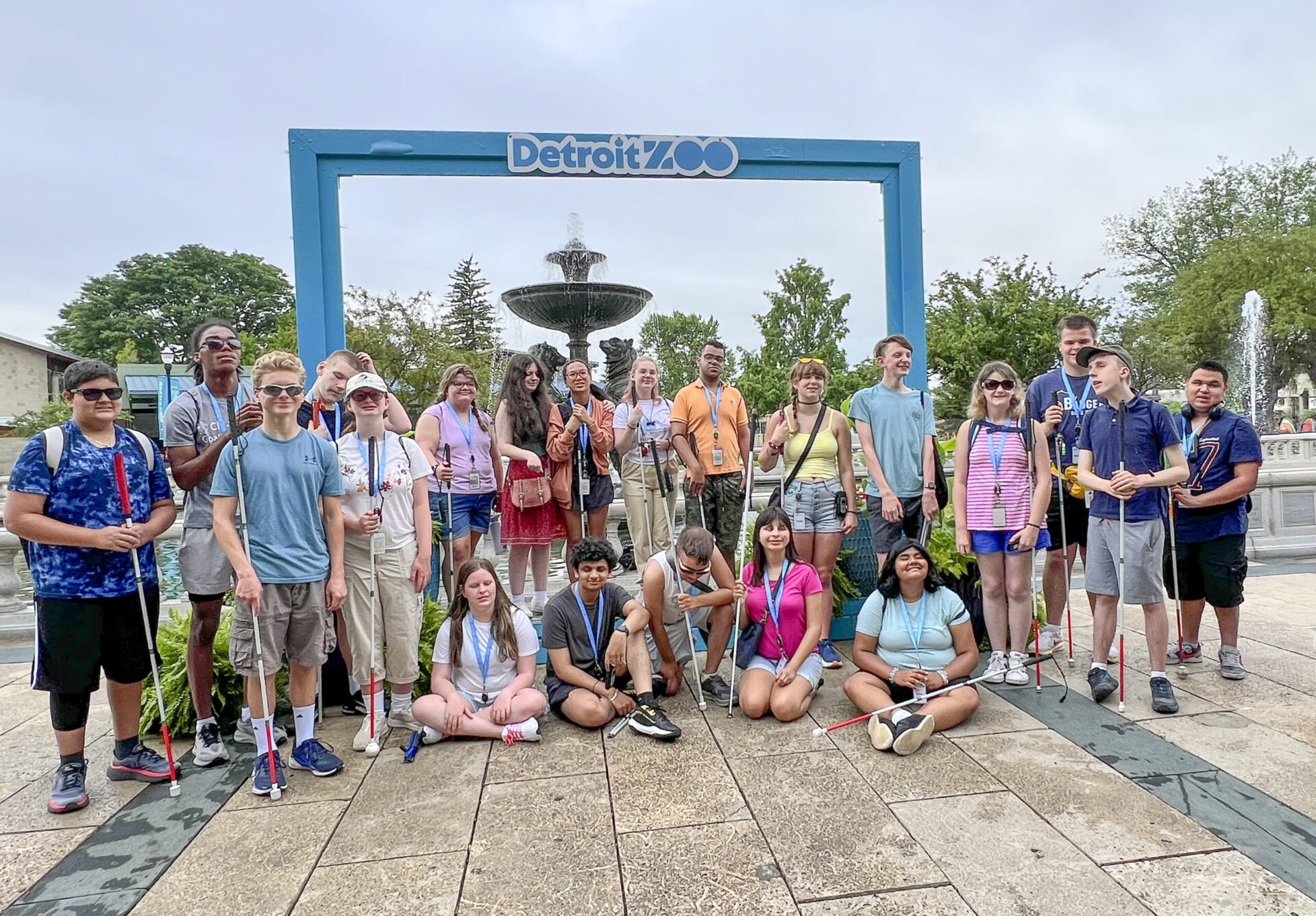A diverse group of individuals poses in front of a large blue sign marking the entrance to the Detroit Zoo, with a fountain and greenery in the background.