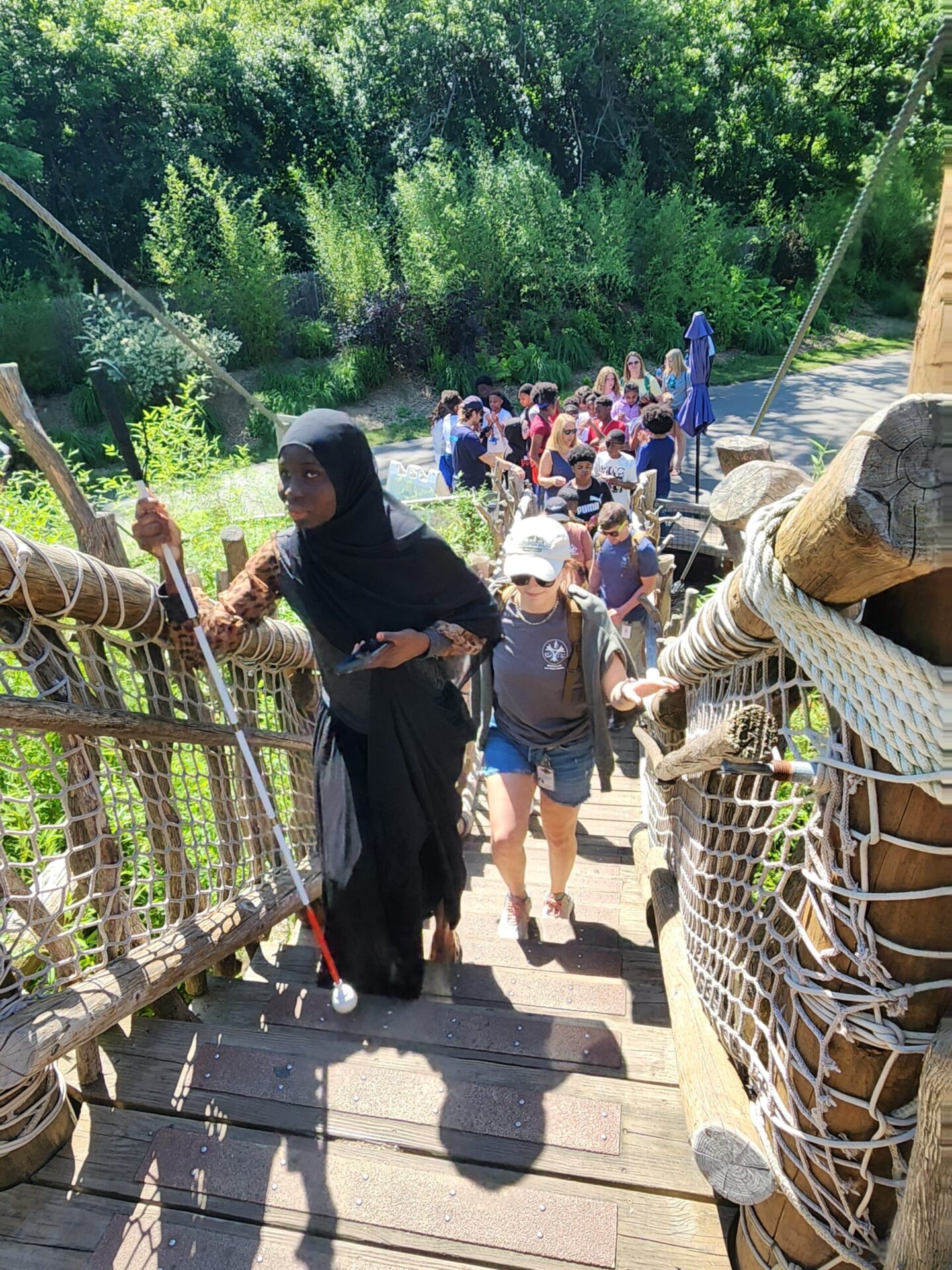 A woman in a black dress uses a white cane while walking up a wooden bridge alongside a group of people in a lush, green setting.