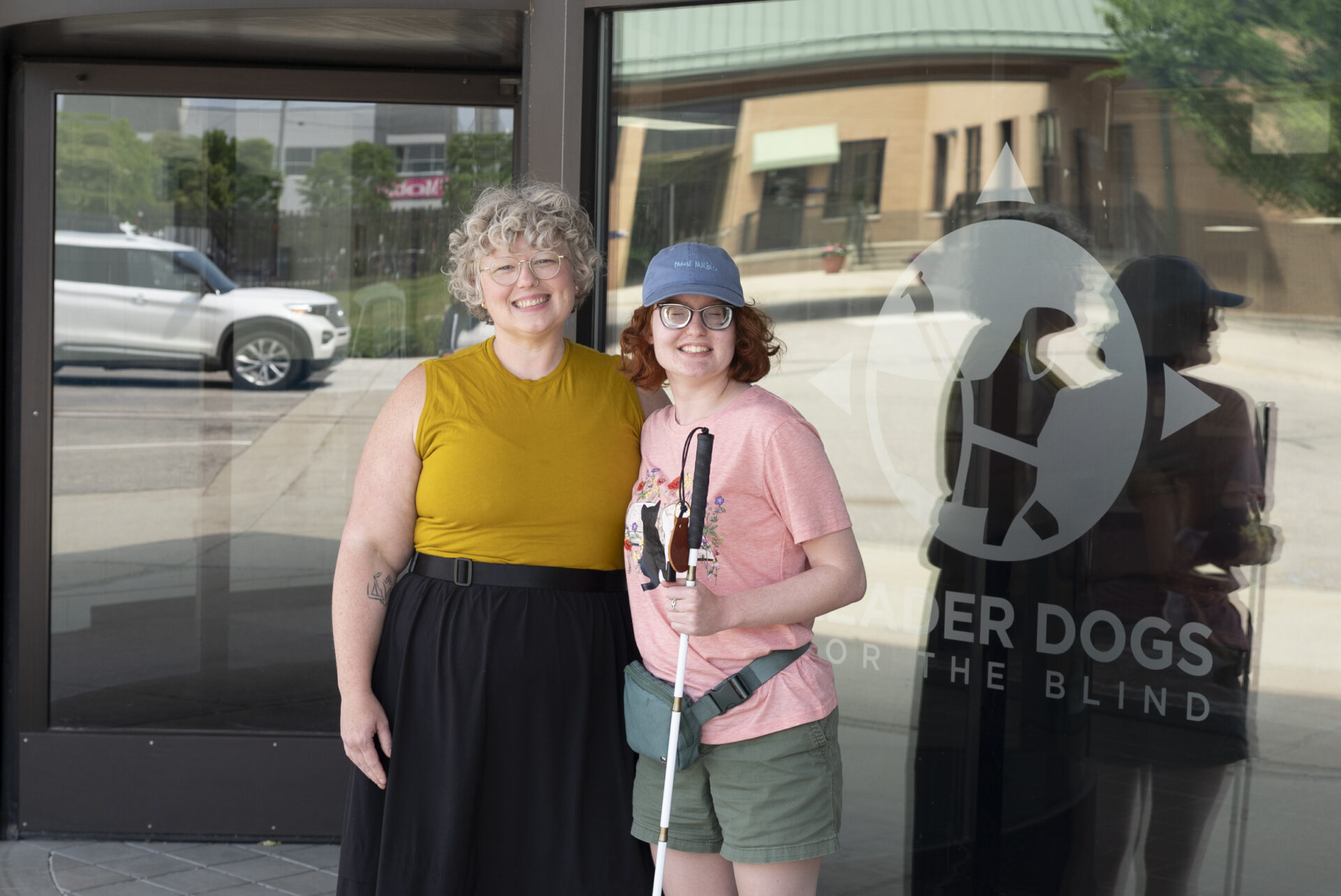 Two smiling women pose in front of a glass door with a Leader Dogs for the Blind logo; one holds a white cane.